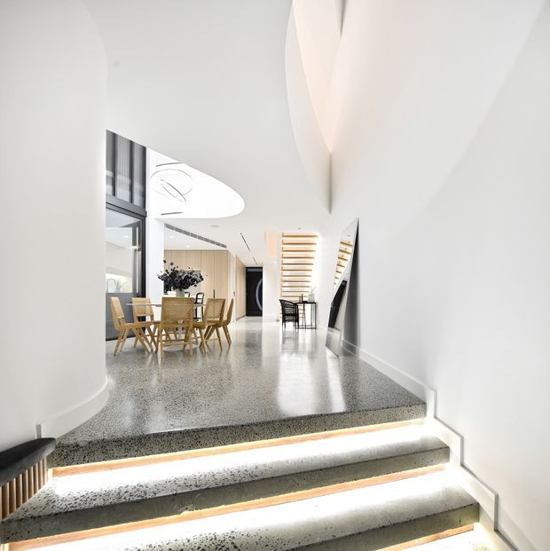 Bright contemporary interior viewed from a short flight of illuminated terrazzo steps, opening into an open-plan dining and circulation area. The space features curved white walls and ceiling forms, polished speckled flooring, full-height glazing, pale timber dining furniture, black accents, and a sculptural floating timber stair screen. Integrated step lighting creates a crisp, refined atmosphere.