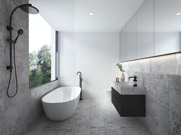 A contemporary bathroom featuring a freestanding white oval bathtub beside a large window, a black wall-mounted rainfall shower, a floating dark vanity with a pale stone countertop, and extensive grey stone-look wall and floor finishes. A large mirrored cabinet spans the vanity wall, with minimalist recessed ceiling lighting and views of greenery outside.