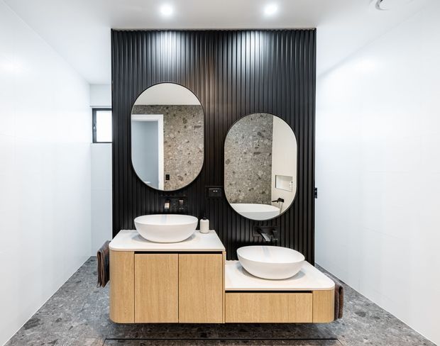 Contemporary bathroom with two vessel basins set on light timber vanities, a dramatic black vertical-ribbed feature wall, large rounded mirrors, white wall finishes, and a mottled grey stone-look floor. The mirrors reflect a textured stone wall and shower area.