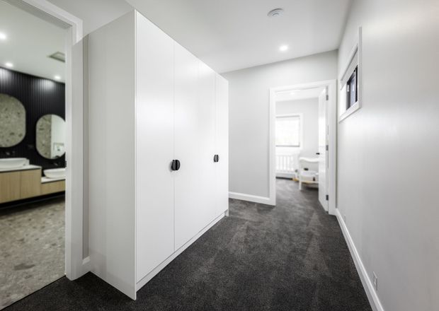 Bright modern hallway with full-height white built-in wardrobes, dark grey carpet, white walls and recessed ceiling lights. An open doorway reveals part of a contemporary bathroom with a dark feature wall and rounded mirrors, while another doorway leads toward a bright bedroom or living area.