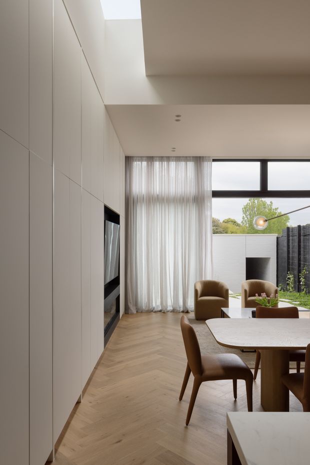 A refined open-plan dining and living area with floor-to-ceiling pale cabinetry concealing a television or fireplace feature on the left. The room has a light timber herringbone floor, soft white sheer curtains, a dark-framed high window, and a sculptural dining table with warm brown upholstered chairs. A small landscaped courtyard is visible beyond the glazing, with a contemporary neutral palette and abundant natural light.