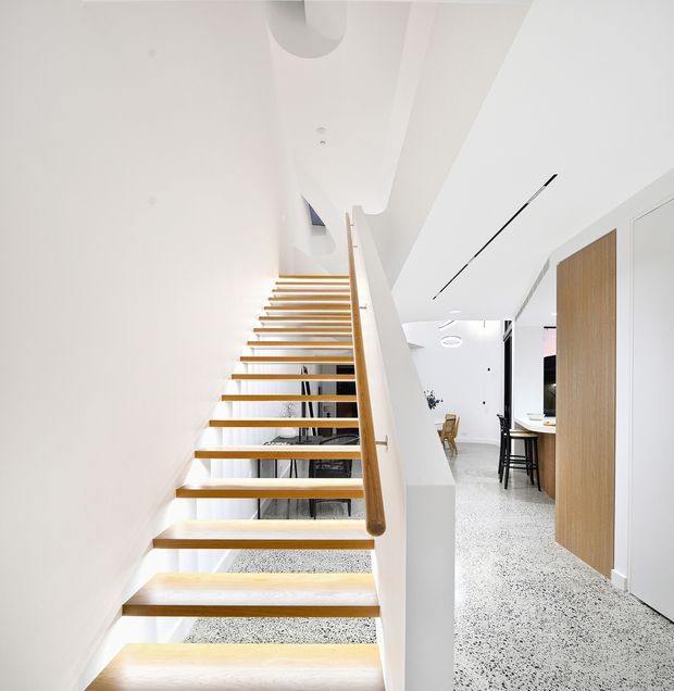 Bright contemporary interior view centered on an open-riser timber staircase with a rounded timber handrail. White walls and ceilings, integrated linear lighting, a pale terrazzo-style floor, and simple white cabinetry create a clean minimalist composition. A dining and kitchen area is visible beyond the stair.