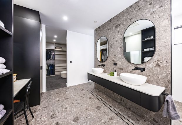 Contemporary bathroom with a double vanity featuring two white vessel basins, matte black cabinetry and tapware, and rounded mirrors mounted on a textured grey stone-look feature wall. Matching grey flooring, integrated shelving, and an open wardrobe area are visible beside the vanity.