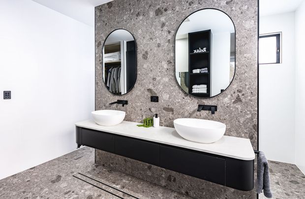 Contemporary bathroom with a double floating vanity, two white vessel basins, black wall-mounted taps, oval mirrors, and a dramatic grey stone feature wall and matching floor. Reflections reveal wardrobe storage beyond the bathroom.