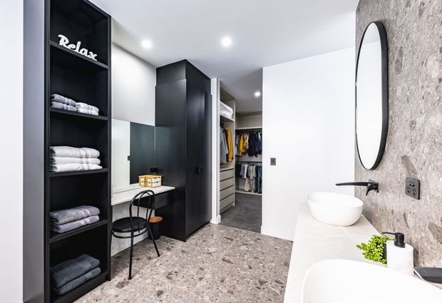 Contemporary bathroom with a freestanding white vessel basin and bathtub, black tapware, large oval mirror, and a textured stone-look feature wall. The room opens directly onto a walk-in wardrobe with dark cabinetry, open shelving, clothing storage, a small desk, and seating. Terrazzo-style flooring and recessed ceiling lights create a clean, modern fit-out.
