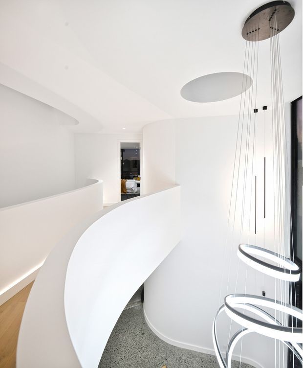 Bright minimalist interior stairway with sweeping curved white balustrades and walls, a polished terrazzo-like floor, warm timber flooring at the landing, and sculptural suspended circular pendant lights extending through the stair void. A dark glazed doorway and tall window add contrast.