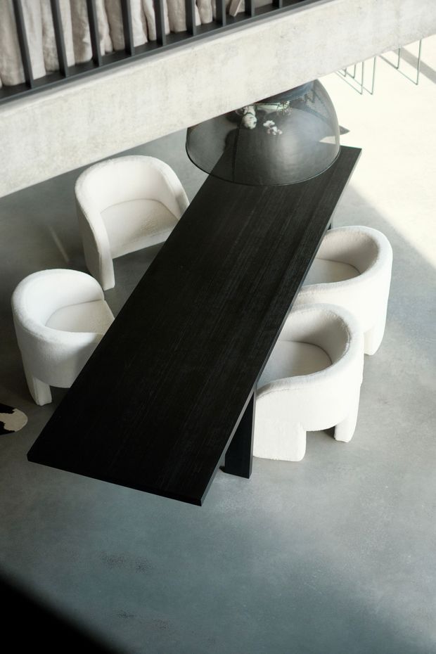 Overhead view of a refined dining area with a long, dark rectangular dining table, four sculptural white upholstered armchairs, and a large smoked-glass dome pendant light. The room features a smooth grey concrete floor, a pale concrete or plaster structural beam, and dark vertical metal railing elements. The high-contrast monochromatic scheme and clean geometric forms create a minimalist, contemporary atmosphere.