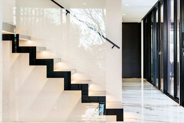 A refined modern interior featuring a sculptural floating staircase with dark, angular stringers and softly illuminated pale treads. A slim black handrail runs along a glossy light wall, while a bright corridor to the right has full-height black-framed glazing and a polished white marble-like floor. Reflections of trees are visible in the glazing and glossy surfaces.