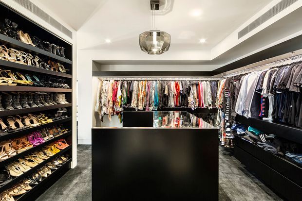 A luxurious walk-in wardrobe with full-height open shelving for shoes and accessories on the left, extensive hanging rails for clothing around the room, and dark built-in cabinetry with a central island. The space has a monochromatic black, white, and grey palette, plush grey carpet, recessed ceiling lighting, linear air-conditioning grilles, and a faceted pendant light.