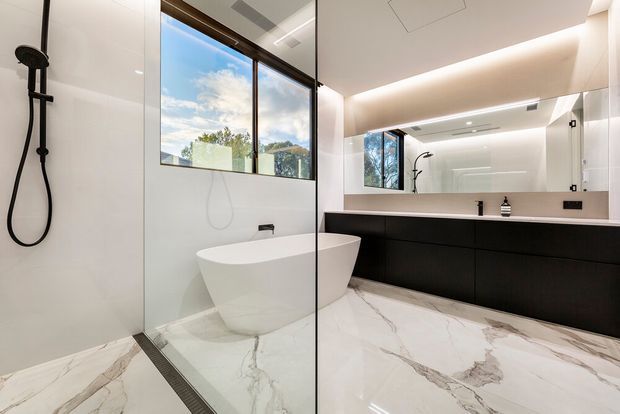 Contemporary luxury bathroom with a freestanding white bathtub positioned beside a frameless glass shower enclosure. The room features large-format white marble-look flooring with grey veining, glossy white wall surfaces, matte black shower and basin hardware, and a long dark vanity with integrated basin. A large mirror and expansive window bring light into the space, creating a bright, refined atmosphere.