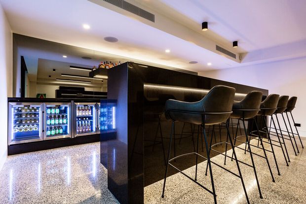 A sophisticated home bar or hospitality-style entertaining area with a long dark feature bar, integrated warm LED strip lighting, and a row of high upholstered bar stools. Backlit glass-door beverage refrigerators and bottle storage are built into the adjacent cabinetry. The polished speckled floor reflects the lighting, while recessed downlights and linear air-conditioning grilles contribute to a sleek, contemporary interior.