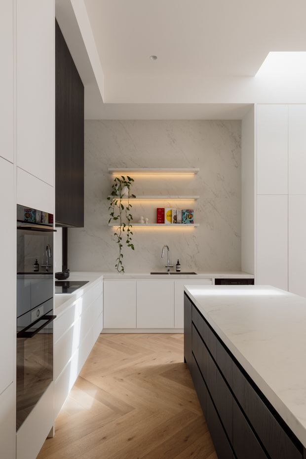 A refined contemporary kitchen with a large central island, integrated white cabinetry, dark timber cabinetry accents, and a pale marble-look splashback. Three floating shelves with warm concealed lighting display books and a trailing plant. The kitchen has a light timber herringbone floor, understated recessed ceiling lighting, and a bright skylight or roof opening.