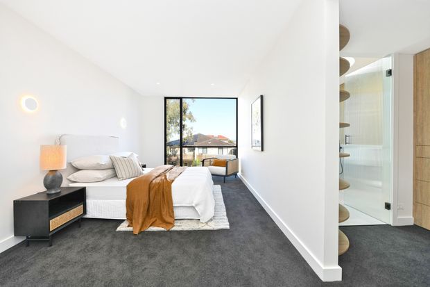 Bright contemporary bedroom with a low-profile upholstered bed, white bedding, rust-coloured throw, dark bedside table, and charcoal carpet. A large black-framed window provides an outlook to neighbouring rooftops and trees, while an open bathroom doorway and curved built-in shelving are visible on the right.