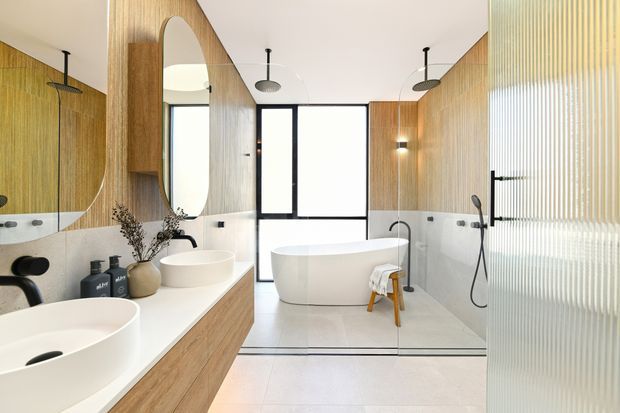Bright contemporary bathroom with a freestanding white bathtub, double vessel basins on a timber vanity, large rounded mirror, black fixtures, and warm vertical timber wall panelling. A translucent ribbed glass shower enclosure occupies the right side, with black-framed glazing at the rear.