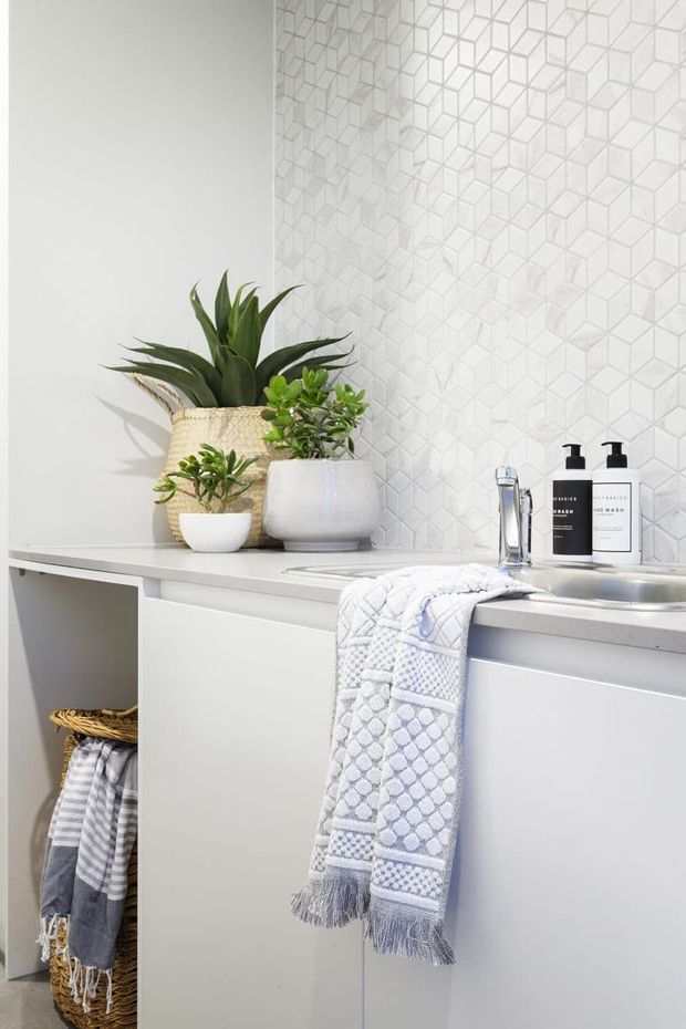 Bright, compact bathroom vanity with a white countertop, integrated basin, pale cabinetry, and a white geometric three-dimensional tile splashback. Potted greenery, soap dispensers, and folded patterned towels add a clean, fresh styling accent.
