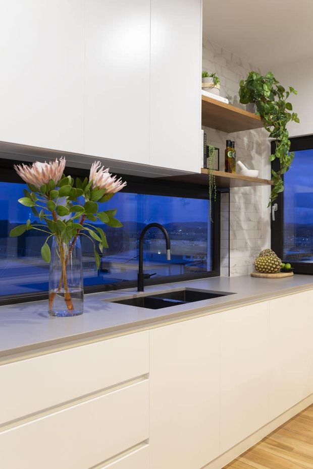 A contemporary kitchen featuring a long white island with integrated cabinetry, a pale solid-surface benchtop, black undermount sink and curved black tap. White overhead cabinetry contrasts with open timber shelving and a light textured tiled splashback. Large dark-framed windows overlook a blue-toned evening cityscape, while plants and flowers add organic detail. Timber flooring is visible at the base.