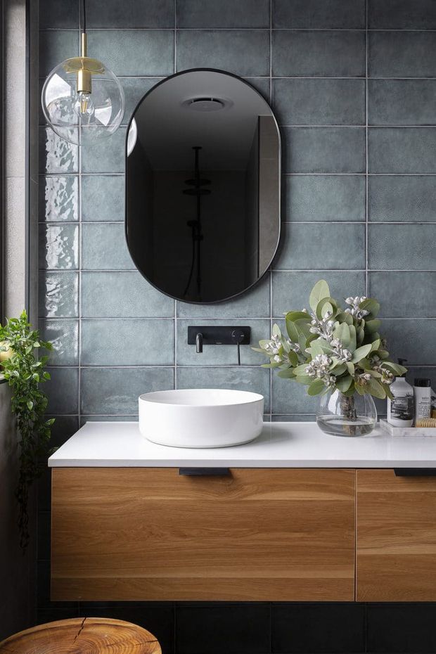 A refined contemporary bathroom vanity viewed straight on. The space features large blue-grey glazed wall tiles, a floating natural timber vanity with a white countertop, a round white vessel basin, matte black wall-mounted tapware, and a black rounded-rectangle mirror. A glass pendant light and leafy floral arrangement add decorative detail.