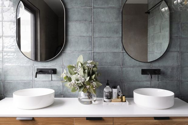 Contemporary double-vanity bathroom with two white vessel basins on a white countertop, dark wall-mounted tapware, grey-green rectangular tiled splashback, rounded black-framed mirrors, and warm timber vanity cabinetry. A floral arrangement and toiletries sit between the basins.