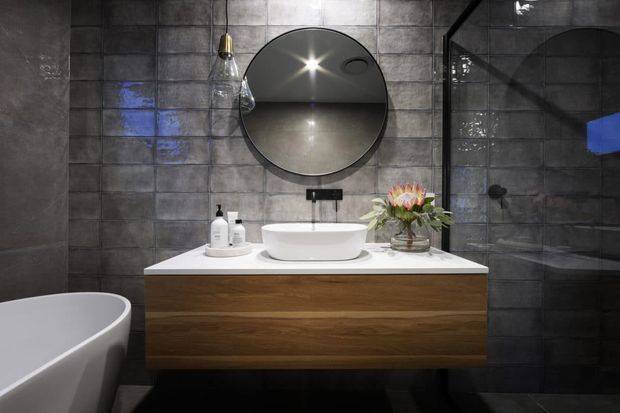 A dark, contemporary bathroom featuring charcoal-grey tiled walls, a floating timber vanity with a white stone benchtop, a white vessel basin, round mirror, black tapware, and a glass-enclosed shower. A freestanding white bathtub is partially visible in the foreground.