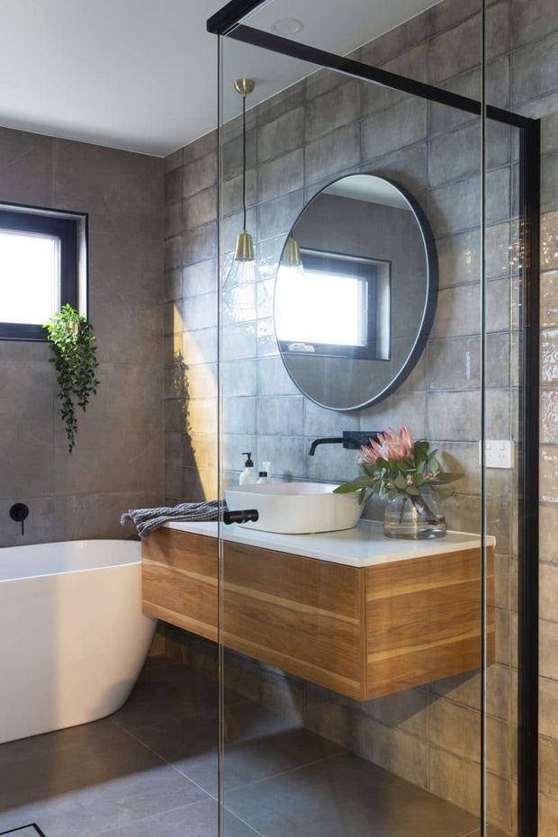 A contemporary bathroom featuring a freestanding white bathtub, frameless glass shower enclosure, floating timber vanity with a white vessel basin, black wall-mounted tapware, a large round mirror, and grey tiled walls and floor. Brass pendant lights and greenery add warmth to the refined space.