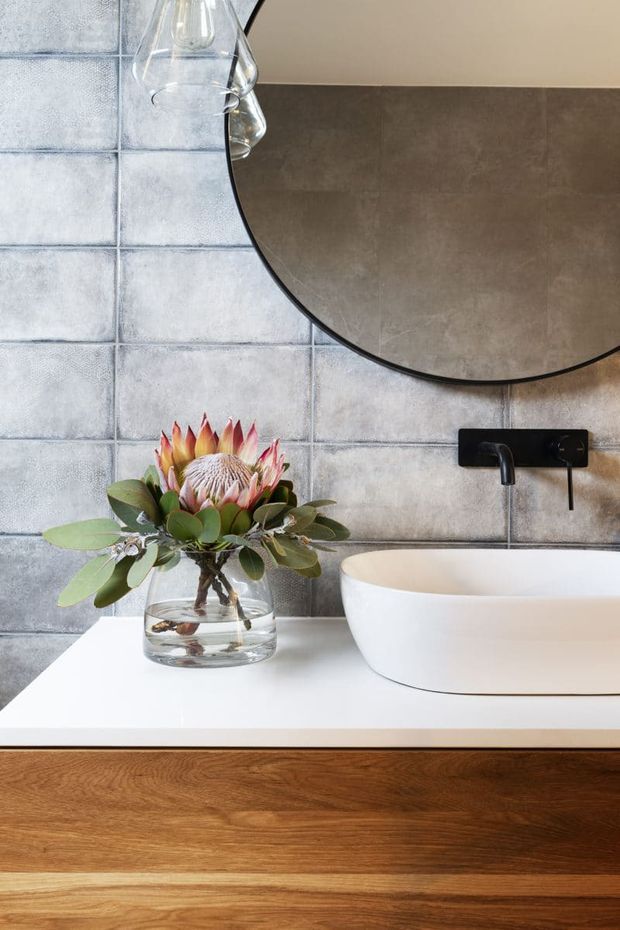 Close-up of a refined contemporary bathroom vanity featuring a white countertop, warm timber cabinetry, a white vessel basin, matte black wall-mounted tapware, large round mirror, and textured pale grey ceramic wall tiles. A glass vase with native flowers adds a natural accent.