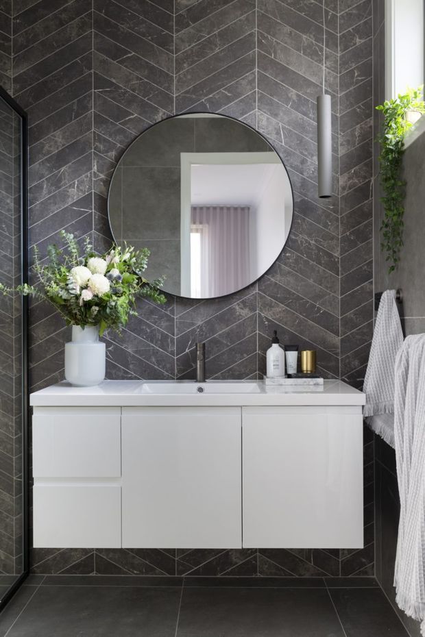 A contemporary bathroom featuring a wall-mounted white vanity with integrated basin, matte black tapware, a large round mirror, and dark grey marble-look herringbone wall tiles continuing onto the floor. A grey towel hangs on the right beside a textured vertical wall feature, while greenery and floral arrangements add a natural accent.