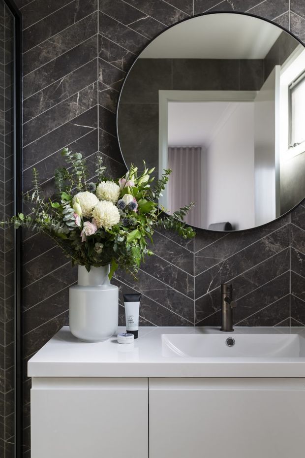 A compact, refined bathroom vanity featuring a white built-in basin and cabinetry, dark marble-look wall tiles in a diagonal herringbone pattern, a round black-framed mirror, and a dark metal tap. A floral arrangement adds an organic accent, while the mirror reflects an adjacent doorway, radiator, and window.