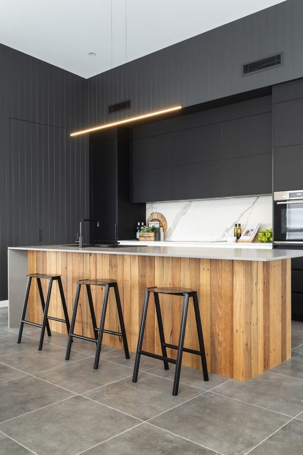 A contemporary kitchen centred on a large island with a warm timber-clad base and pale stone benchtop. Four black bar stools line the island, while dark charcoal vertical wall panelling, matte black cabinetry, integrated appliances, and a white veined splashback create a high-contrast modern interior. A slim linear pendant light illuminates the island, and large grey floor tiles complete the space.