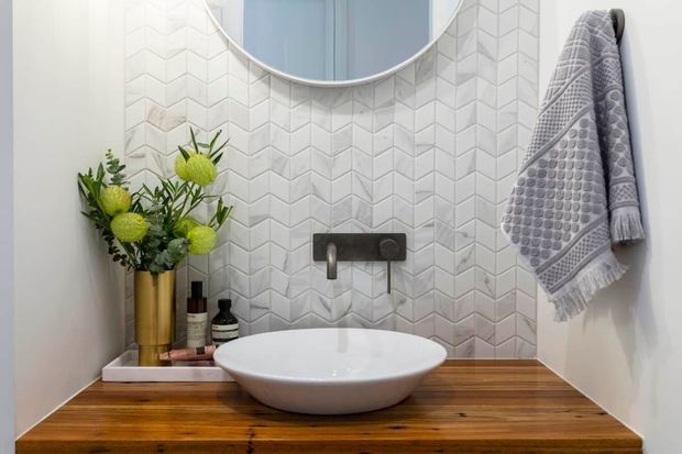 A compact, refined bathroom vanity with a white vessel basin set on a warm timber benchtop. The wall behind features a white geometric chevron tile splashback, a wall-mounted brushed metal tap, and a round mirror. A grey textured towel and decorative greenery add soft contrast.