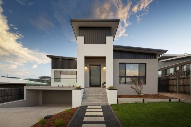 Front exterior view of a contemporary multi-level residence with a strong central entry tower, flat projecting rooflines, white rendered walls, dark vertical cladding, large black-framed windows, and a covered carport or garage area. A landscaped front lawn and stepping-stone path lead to the recessed entrance.