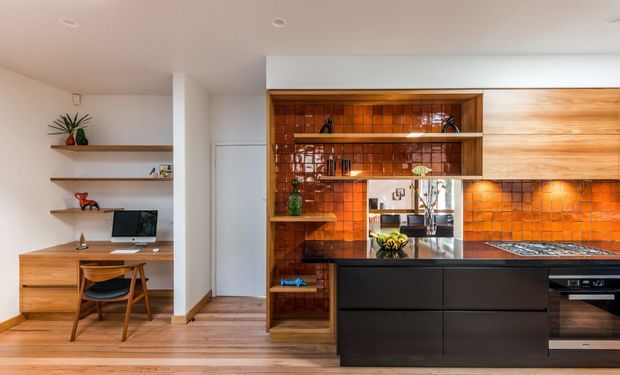 Warm contemporary kitchen and integrated study nook. The kitchen features dark matte cabinetry, a central island with a dark stone benchtop, glossy orange-brown square-tile splashback, open timber shelving, and timber upper cabinetry. An adjacent built-in timber desk with open shelving occupies the left side, while light timber flooring and white walls create a balanced, inviting interior.