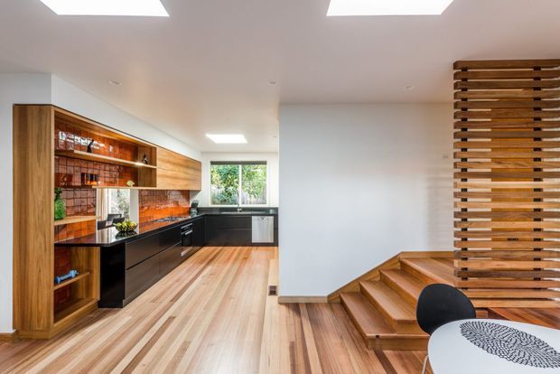 Contemporary open-plan kitchen and stair area with warm timber flooring, extensive timber cabinetry and shelving, dark kitchen joinery, a tiled or patterned splashback, and a sculptural timber-slat screen beside the staircase. Skylights and a rear window provide bright natural light, while a small dining table and chair are visible in the foreground.