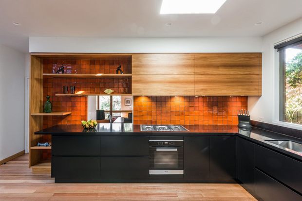 Contemporary kitchen with a large black island and black lower cabinetry, warm timber overhead cabinetry and open shelving, and a striking orange-red tiled splashback. Integrated cooktop and oven, dark benchtops, recessed lighting, a skylight, and a large window create a refined, warm, high-contrast interior.