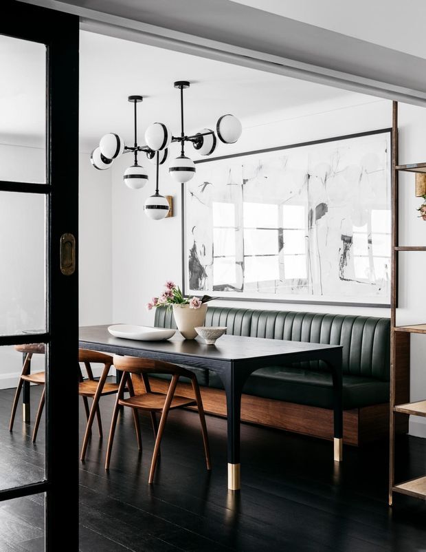 A refined dining area viewed through black-framed glazed doors, featuring a long dark dining table with timber chairs, a built-in channel-tufted dark green banquette, black timber flooring, a large abstract artwork, open timber shelving, and a sculptural multi-globe pendant light. The room has a sophisticated, high-contrast contemporary look with warm timber accents.