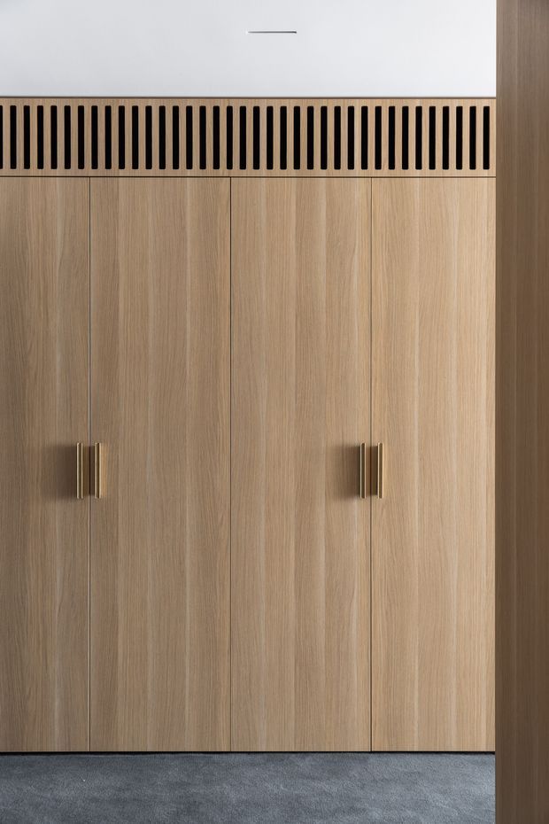 Close-up view of a minimalist built-in timber wardrobe or storage wall. The tall vertical cabinet doors have a light natural wood-grain finish and slim brass-toned handles. A black vertical slatted ventilation grille runs beneath a white upper panel, with dark grey carpet flooring below.