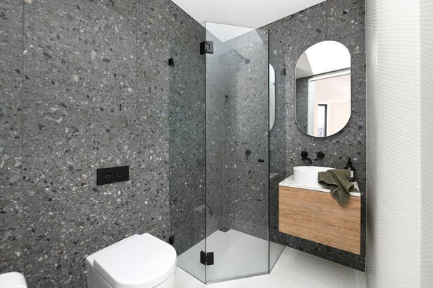 Contemporary bathroom featuring dark grey terrazzo wall and floor finishes, a frameless glass corner shower, wall-hung toilet, and a floating timber vanity with a white vessel basin. A rounded-rectangle mirror and matte black fittings add contrast.