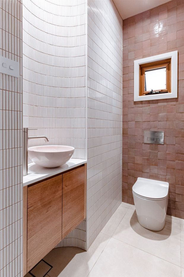 Compact contemporary bathroom with a curved wall of narrow white tiles, pale tiled flooring, a wall-hung timber vanity with a white vessel basin, brushed metal tapware, and a toilet. The opposing wall is finished in glossy dusty-pink rectangular tiles and includes a small timber-framed window with white trim and a concealed cistern flush plate.