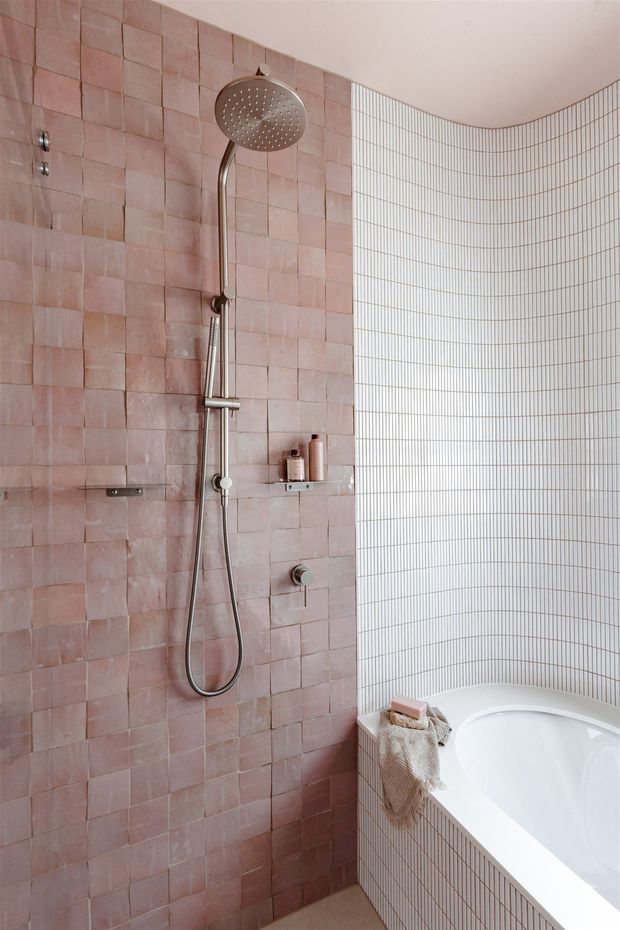 A contemporary bathroom featuring a walk-in shower with brushed metal rainfall showerhead and handheld fitting. The shower walls are clad in textured dusty pink square tiles, contrasting with a curved wall and bathtub surround finished in narrow white grid tiles. A white freestanding or integrated tub is partially visible, with minimal bath accessories and a warm, refined palette.