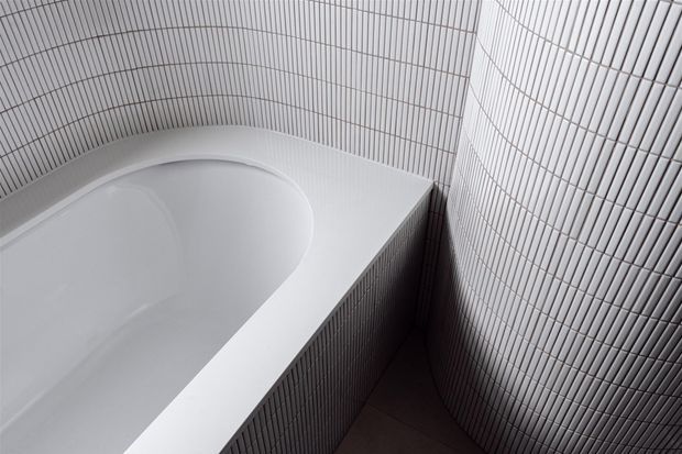 Close-up of a minimalist bathroom corner featuring a white freestanding or built-in bathtub surrounded by vertically ribbed white wall and bath-panel tiles. The monochromatic surfaces create a clean, sculptural and highly textured composition.