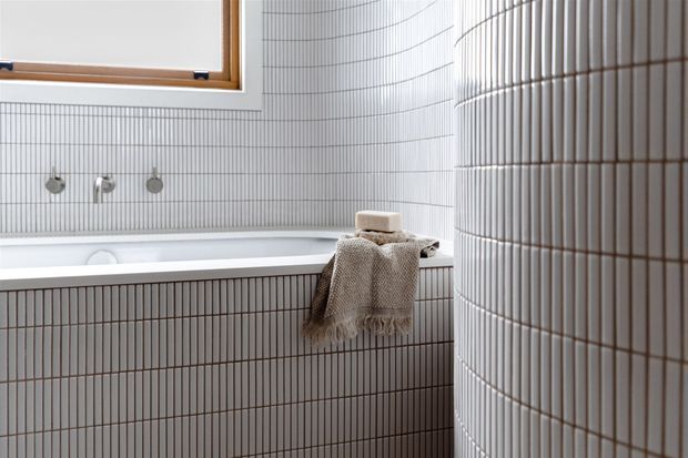 A bright, refined bathroom featuring a built-in bathtub enclosed by vertically ribbed, narrow white tiles. Matching tiled walls and curved tiled partition create a softly sculptural effect, complemented by brushed metal wall-mounted tapware, a timber-framed window, and a neutral textured towel.