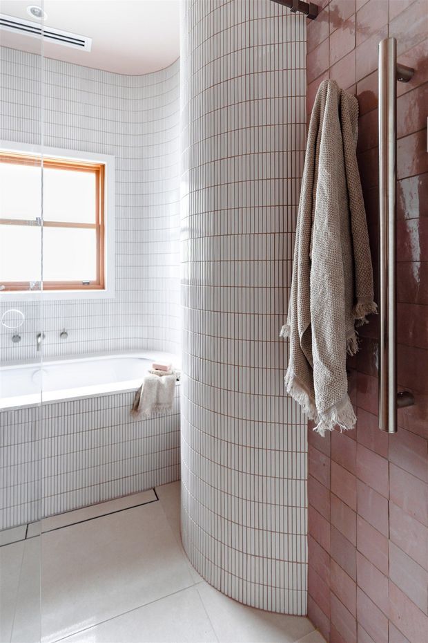Bright contemporary bathroom featuring a freestanding bathtub set against curved walls clad in fine white vertical mosaic tiles. A curved tiled partition separates the bath from a shower area with dusty pink rectangular wall tiles, a brushed metal grab rail, and a neutral textured towel. Timber-framed window and pale stone-look floor add warmth and light.