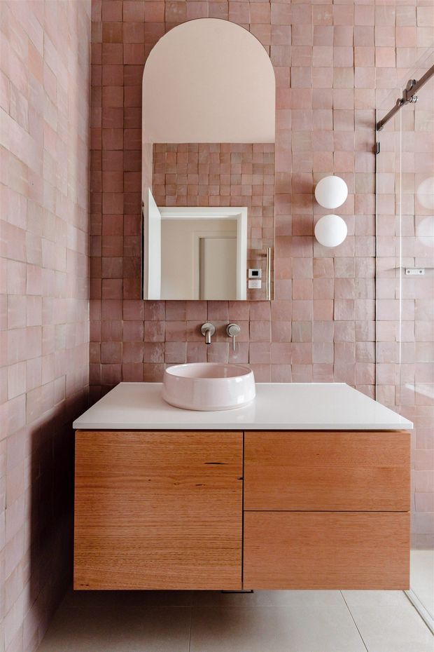 A compact contemporary bathroom featuring blush pink square wall tiles, a floating natural timber vanity with a white benchtop, a round vessel basin, wall-mounted brushed metal taps, and an arched mirror. A glass shower enclosure and spherical wall lights are visible on the right.