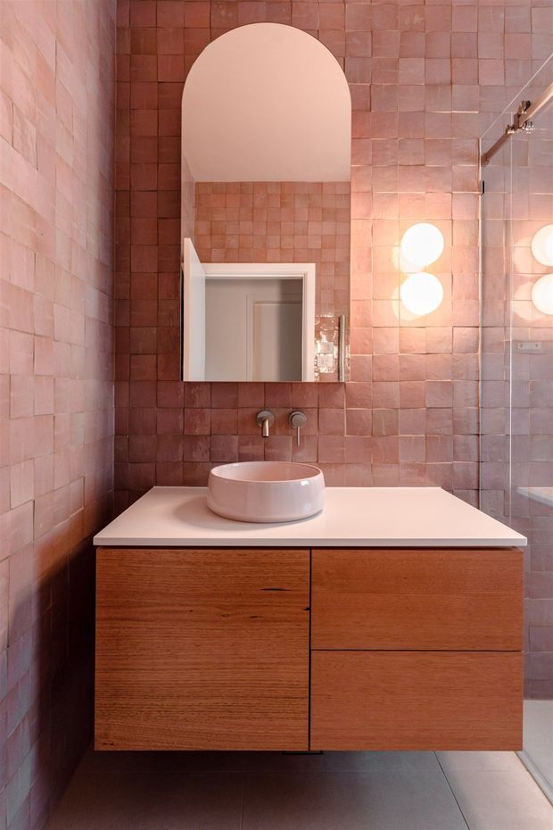 A compact contemporary bathroom with dusty pink square wall tiles, a wall-mounted warm timber vanity, pale solid-surface benchtop, round vessel basin, wall-mounted tapware, and an arched mirror. A glass shower enclosure and round wall lights are visible on the right.