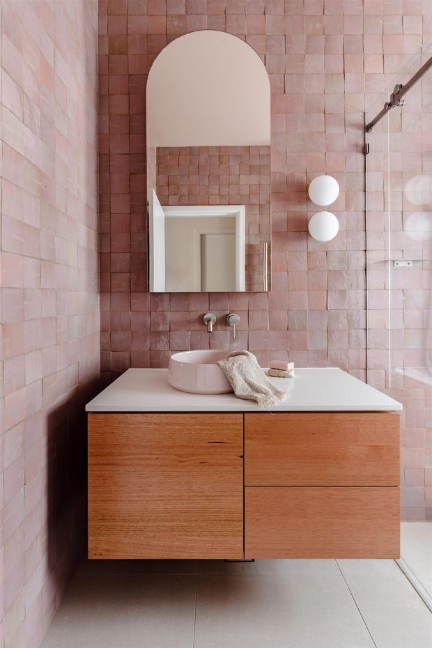 A refined contemporary bathroom featuring dusty pink square wall tiles, a floating natural timber vanity with a white benchtop, integrated wall-mounted tapware, a pale pink vessel basin, an arched mirror, and globe wall lights. Large neutral floor tiles and a frameless glass shower enclosure complete the calm, tonal scheme.