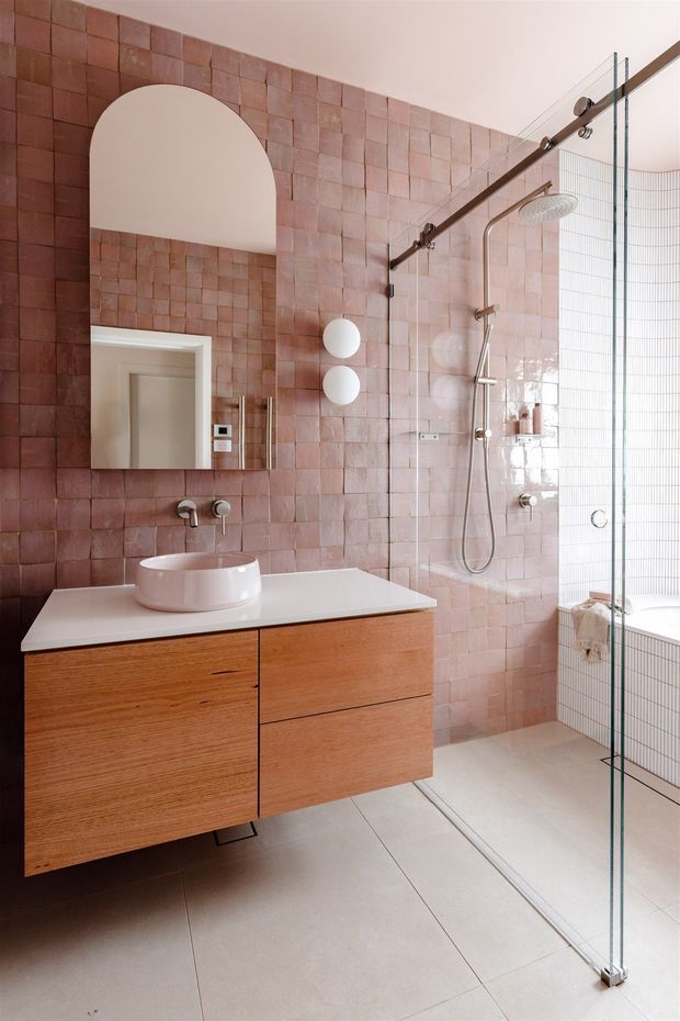 Contemporary bathroom featuring dusty pink square wall tiles, large pale floor tiles, a floating natural timber vanity with white benchtop and vessel basin, and a frameless glass walk-in shower. A freestanding or built-in tub is partially visible behind the shower glazing, with a curved recessed wall niche and brushed metal fixtures.