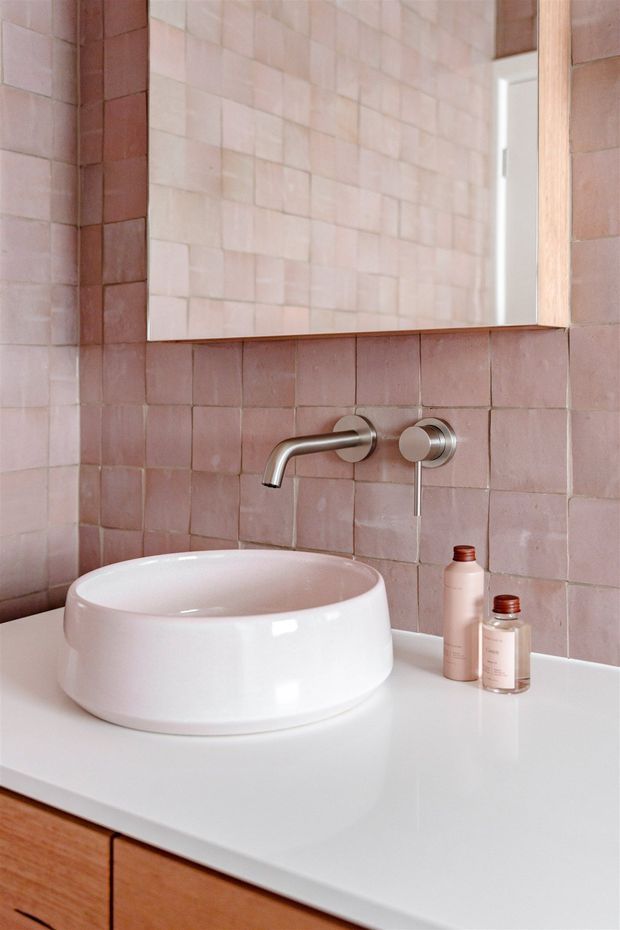 A close-up view of a refined bathroom vanity featuring a white vessel basin on a white benchtop, wall-mounted brushed metal tapware, and warm timber cabinetry. The walls are clad in blush-pink square tiles, with a large frameless mirror above the basin and matching neutral-toned toiletries.