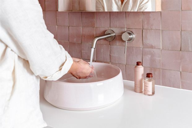 A close-up bathroom vanity scene with a round white vessel basin on a white benchtop, wall-mounted chrome tapware, and dusty pink square tiled splashback. A person in a white robe washes their hands, with matching pink-toned bottles beside the basin and a mirror above.