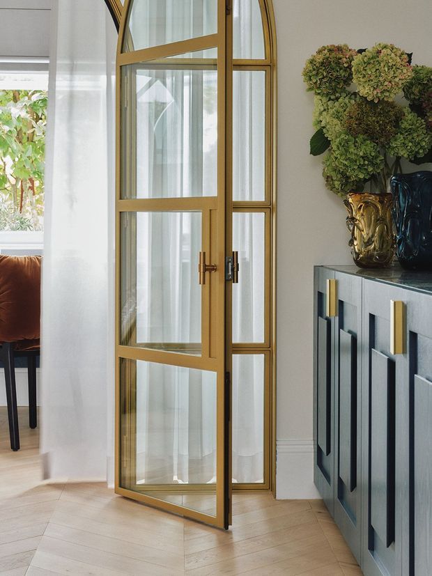 Refined interior detail featuring a tall glazed timber-framed door with warm brass-gold finish and decorative arched framing. The door opens beside a dark blue-grey built-in cabinet with brass hardware, while sheer curtains, pale timber flooring, and a floral arrangement create an elegant, softly lit atmosphere.