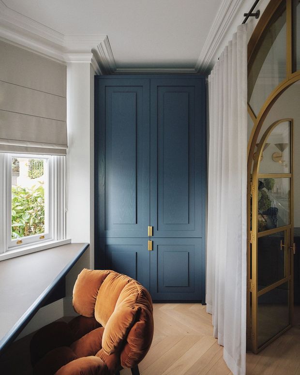 Refined interior corner featuring a deep navy-blue paneled built-in cupboard, white walls with elaborate crown moulding, a light herringbone timber floor, and a window with a Roman blind. A sculptural rust-orange velvet armchair sits beside a dark built-in desk or console. On the right, fluted white curtains frame a distinctive arched, bronze-toned glazed door or cabinet. The space has a tailored, elegant and softly layered character.