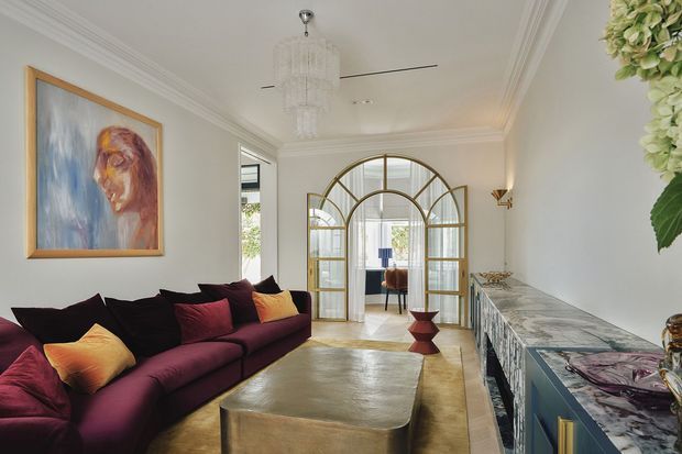 Elegant living room with a deep burgundy sectional sofa, jewel-toned cushions, a large metallic coffee table, and a richly patterned marble-look built-in sideboard. The room features white decorative cornices, a crystal chandelier, brass-framed arched glass doors, wall sconces, timber flooring, and a colourful framed artwork, creating a refined, glamorous interior.